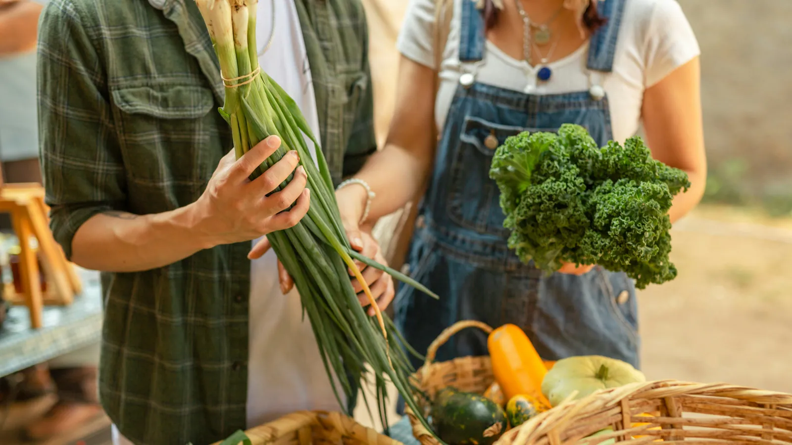 Deux personnes tiennent des oignons verts et du chou frisé près de paniers de courges à un kiosque de marché