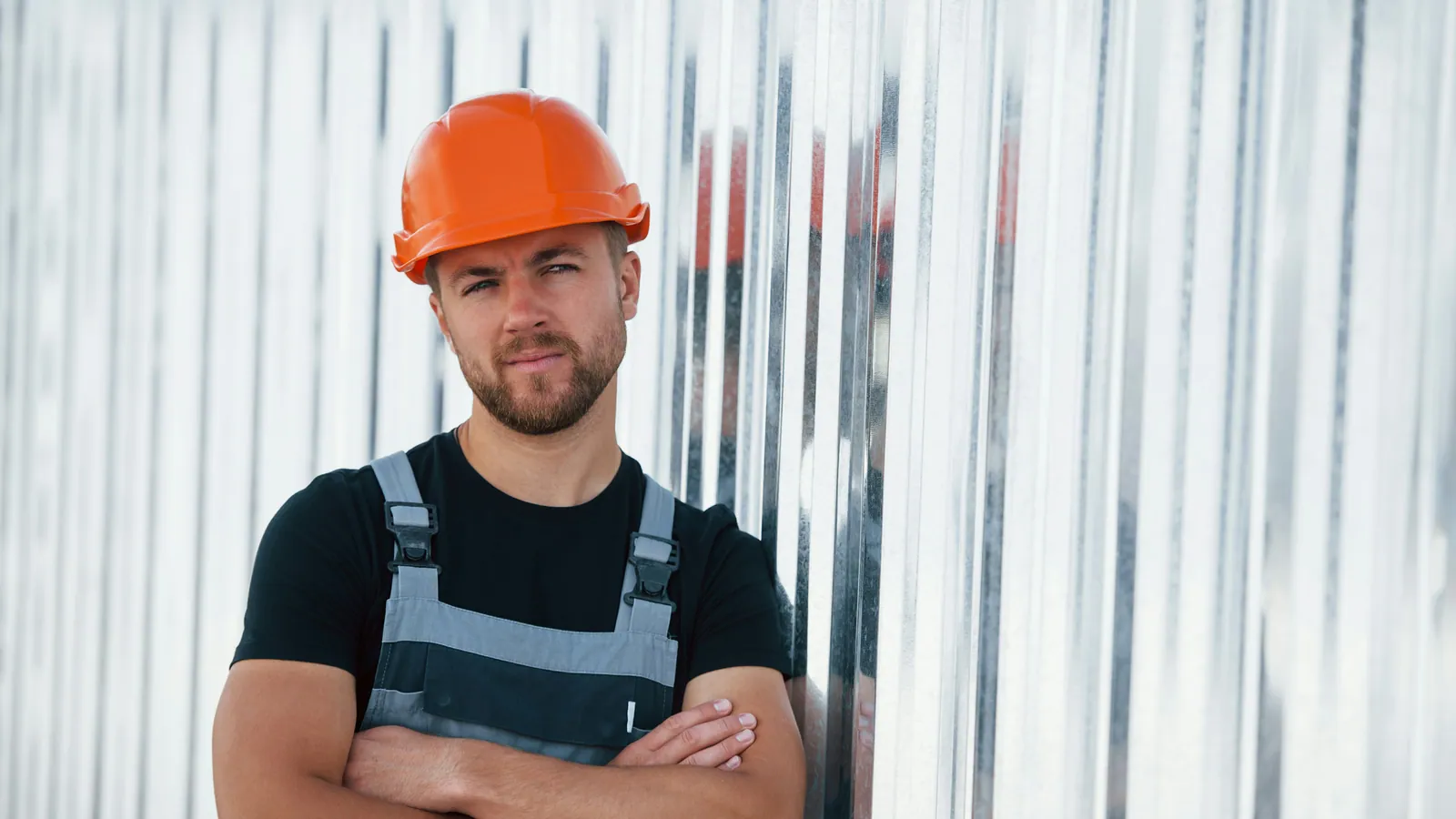 Travailleur avec casque de sécurité orange et salopette grise, bras croisés devant un mur de tôle ondulée