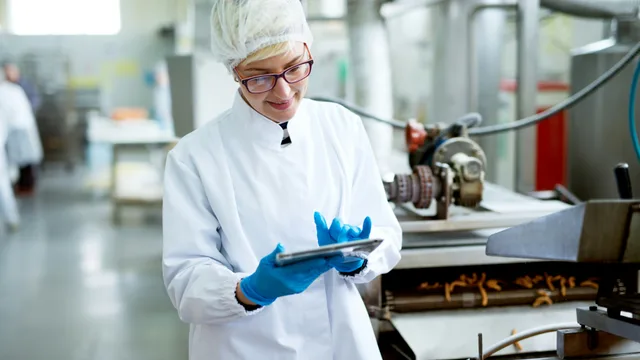 Agri-food plant employee in blue gloves and a hairnet holding a tablet beside a snack production line