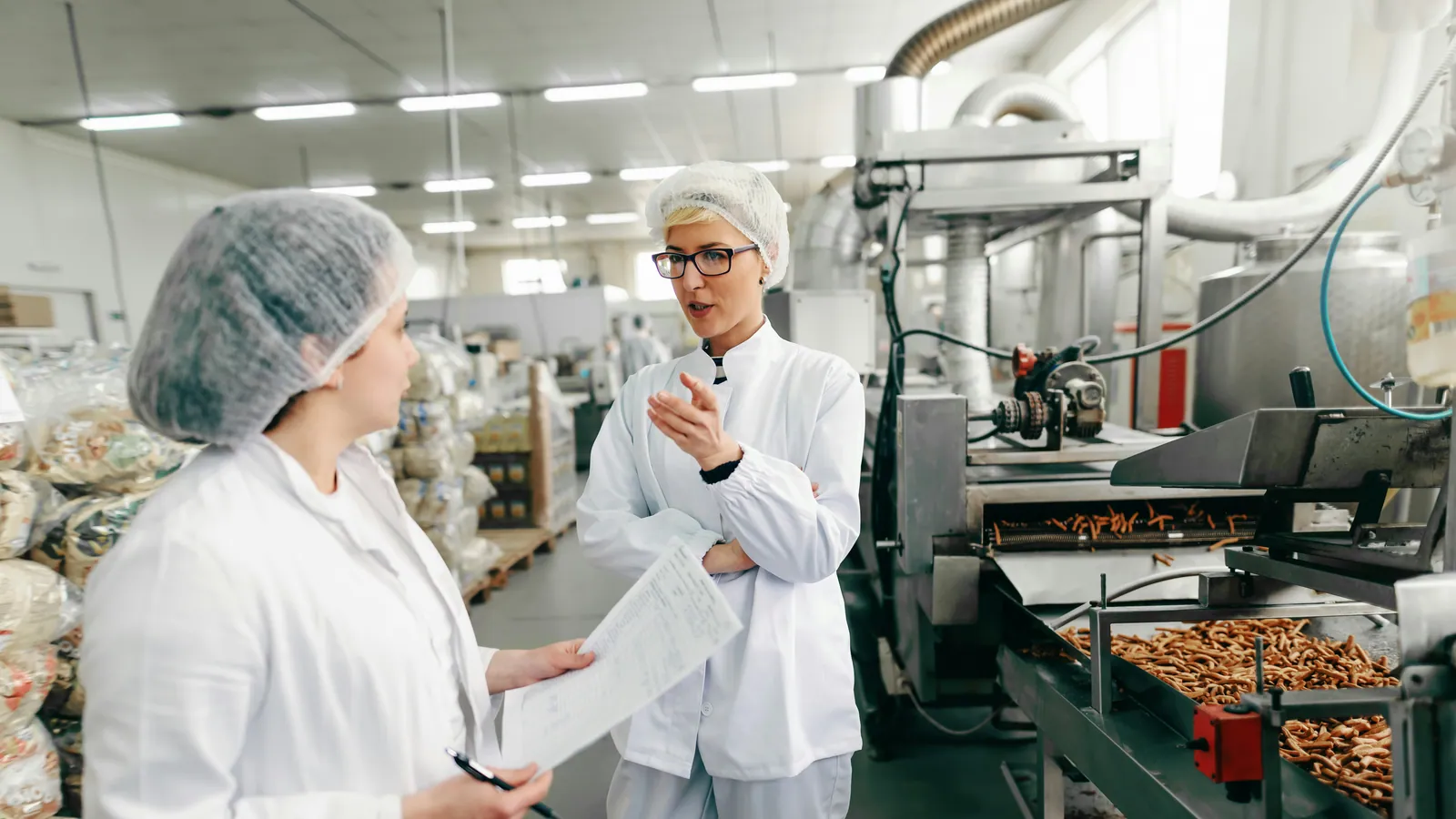 Employees in lab coats discussing a document beside a snack production line, a project type covered by food processing grants